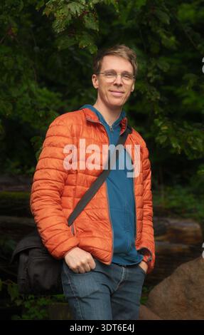 Selbstbewusster, lässiger junger Mann. Außenporträt eines glücklichen kaukasischen männlichen Erwachsenen, der in der Natur eine orange Jacke und eine Tasche trägt. Stockfoto