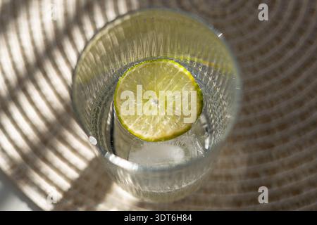 Über dem Kopf lag ein klares Glas, gefüllt mit gekühltem Wasser, frischen Limettenscheiben und Eiswürfeln, vor einem sauberen weißen Hintergrund, warmes Licht Stockfoto