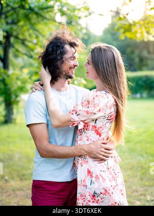 Glückliches Paar, das in einem sonnigen Park nahe beieinander steht, lächelt und Hände hält Stockfoto