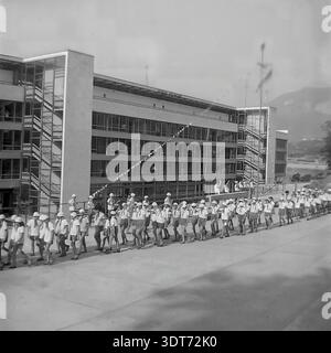 Ein historisches Schwarzweiß-Foto aus den 1960er Jahren, das eine lange Kolonne sowjetischer Kinder zeigt, die über einen geräumigen Platz im berühmten All-Union Artek Pionierlager in Gurzuf auf der Krim marschieren. Jungen und Mädchen in traditionellen Sommeruniformen mit panamahüten und Krawatten treten gemeinsam auf. Im Hintergrund zeigt ein modernes, mehrstöckiges Wohnheim mit weitläufigen Verglasungen und offenen Treppen die Architektur des Resorts. Dieses Archivbild zeigt organisierte Sommererholung, Disziplin und eine friedliche Kindheit in der UdSSR. Stockfoto