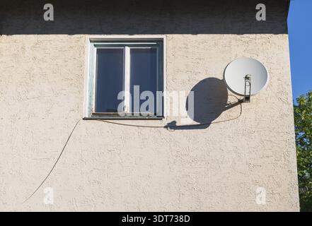 Eine weiße Satellitenschüssel auf einer beigefarbenen Stuckwand neben einem alten Fenster mit weißem Rahmen. Blauer Himmel und grüne Bäume im Hintergrund. Kabel verlaufen von Stockfoto