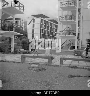 Ein historisches Schwarzweiß-Archivfoto aus den 1960er Jahren, das die beeindruckende modernistische Architektur eines mehrstöckigen Schlafsaals im legendären All-Union Artek Pionierlager in Gurzuf auf der Krim feststellt. Die Szene zeigt Frauen und Kinder, die auf einer langen Bank in einem gepflasterten Innenhof ruhen, mit der innovativen Struktur mit Außentreppen und zahlreichen Balkonen im Hintergrund. Umgeben von üppigem Grün und der malerischen Landschaft der Krim veranschaulicht dieses Bild die friedliche Atmosphäre organisierter Sommerferien in der UdSSR. Stockfoto
