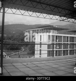 Ein historisches Schwarzweiß-Archivfoto aus den 1960er Jahren, das einen malerischen Blick von einer erhöhten Terrasse im berühmten All-Union Artek Pionierlager in Gurzuf auf der Krim bietet. Das Bild zeigt moderne, mehrstöckige Schlafsäle mit Flachdächern und großen Fenstern, die die moderne sowjetische Architektur der damaligen Zeit darstellen. Die nebligen Krim-Berge bilden eine dramatische Kulisse hinter dem üppigen Grün und dem gepflasterten Innenhof. Diese eindrucksvolle Szene bewahrt die visuelle Erinnerung an das Leben vor dem Krieg und zeigt das große Ausmaß der Resortentwicklung und die friedliche Atmosphäre der Sommerferien in der UdSSR. Stockfoto
