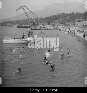 Auf diesem Schwarzweiß-Archivfoto aus den 1960er Jahren entfaltet sich am Strand von Gurzuf auf der Krim eine lebhafte Sommerszene. Zahlreiche Urlauber schwimmen im ruhigen Schwarzen Meer, während in der Nähe ein weißes Rettungs-Motorboot mit dem Wort Spasatelny steht und drei Männer an Deck stehen. Im Hintergrund ragt ein massiver Industrieportalkran über dem Hafengebiet vor den malerischen Küstenhügeln. Dieses ehrliche Bild fängt die einzigartige Atmosphäre des sowjetischen Resortlebens ein und verbindet Küstenerholung mit industriellem Fortschritt in der Mitte des 20. Jahrhunderts. Stockfoto