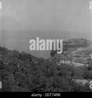 Vintage-Schwarz-weiß-Archivfoto, das die berühmte Burg Swallow's Nest in den 1960er Jahren auf der Aurora Cliff in Gaspra auf der Krim zeigt Die malerische Aussicht überblickt das ruhige Schwarze Meer, mit einem bewaldeten Hügel im Vordergrund und Zeichen von Bautätigkeit mit aufsteigendem Rauch, der unter der Klippe sichtbar ist. Dieses wunderschön erhaltene historische Bild spiegelt perfekt das architektonische Erbe und die Küstenschönheit der Halbinsel Krim während der Sowjetzeit wider. Stockfoto