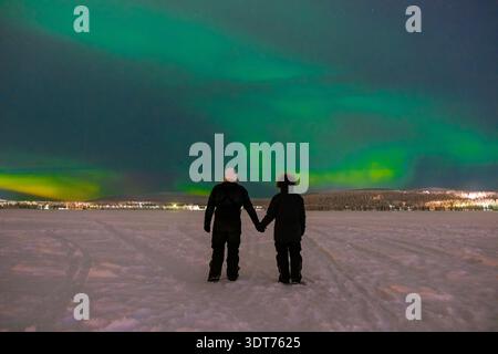 Ein Paar hält Hände auf einer schneebedeckten Landschaft, fasziniert von den leuchtenden Nordlichtern, die am Nachthimmel tanzen. Akaslompolo, Lappland Finnland Stockfoto