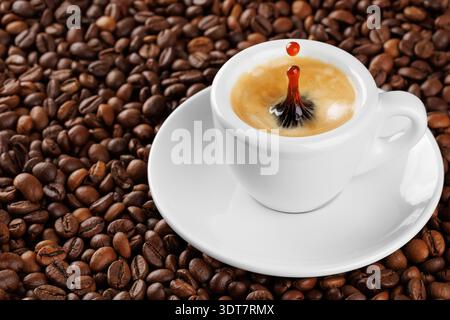 Kleine weiße Espresso-Tasse mit Spritzern und Tropfen, voller aromatischem Kaffee, der auf einem Bett aus gerösteten Bohnen liegt Stockfoto