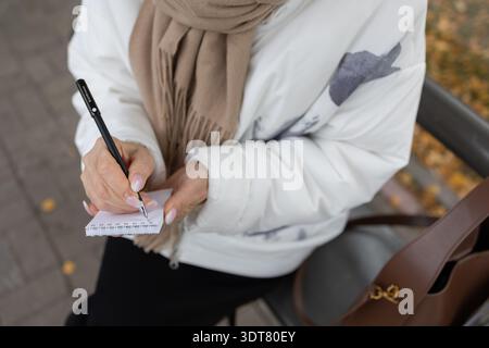Durchdachte Frau in weißer Jacke und beigefarbenem Schal schreibt Notizen, während sie im Herbst an der Metallbank steht, Hände in Nahaufnahme Stockfoto