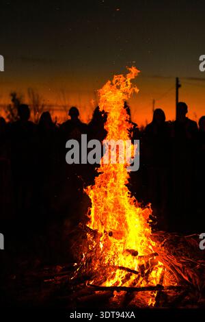 Vertikales Foto. Ein massives Lagerfeuer sendet Flammen nach oben, während sich eine dichte Gruppe von Silhouetten versammelt, die von feurigem Licht beleuchtet werden. Das Feuer steigt in den auf Stockfoto