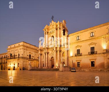Die Aussicht zeigt die Kathedrale von Syrakus, die sich in der Abenddämmerung auf der Insel Ortygia auf Sizilien befindet. Das Gebäude steht hoch mit Lichtern, die seine Fassade beleuchten. Stockfoto