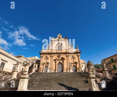 Die Kirche Chiesa di San Pietro steht hoch in Modica, Sizilien. Das starke Sonnenlicht scheint auf die Stufen der Kirche, während ein klarer blauer Himmel die Kulisse erfüllt. Stockfoto