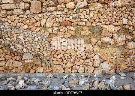 Hochauflösende Aufnahme einer alten rustikalen Steinmauer aus natürlichem Felsen, der auf einer Sandoberfläche mit verstreuten Kieselsteinen und organischen Abfällen steht. Stockfoto
