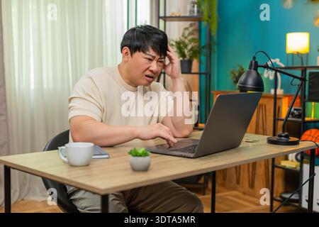 Asiatischer Mann, der am Tisch sitzt, liest schlechte E-Mails auf dem Laptop erneut, klopft wütend auf die Tastatur, die Stirn tief gefurcht. Der Chinese zeigt eine angespannte Frustration, Shou Stockfoto
