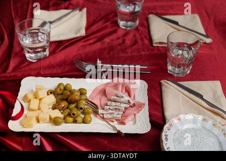 Ein Tisch auf rotem Samttuch ist mit einer weißen rechteckigen Platte mit Käsewürfeln, gemischten grünen Oliven, gefalteten Mortadella-Scheiben und ein paar Pers Stockfoto