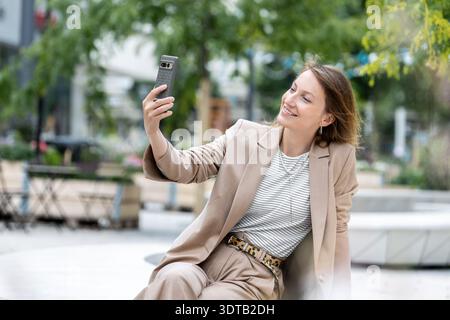 Lächelnde Geschäftsfrau trägt beigefarbenen Anzug Nehmen Sie Selfie Smartphone Urban Park Genießen Sie die Sommerpause. Moderner Lifestyle Portrait Selbstvertrauen Eleganz Stockfoto