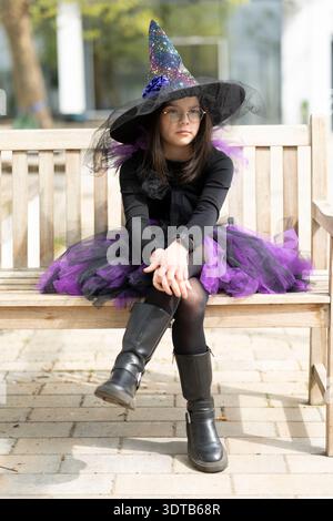 Ein nachdenkliches Mädchen in Brille, das ein Hexenkostüm trägt, sitzt auf einer Holzbank, sichtbare Lederstiefel, blickt in die Ferne. Konzept von Halloween, Stockfoto