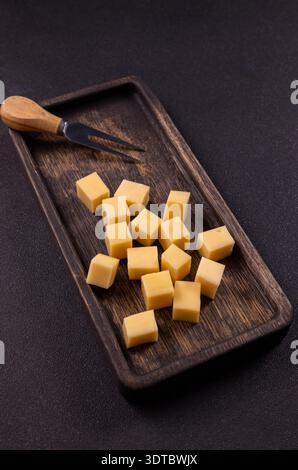 Käsewürfel und eine Vorspeisengabel ruhen auf einem dunklen rustikalen Holztablett und schaffen eine einfache und elegante Snackpräsentation auf einem strukturierten Backgr Stockfoto