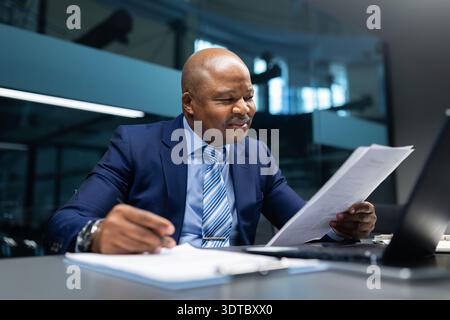Ein lächelnder schwarzer Geschäftsmann liest am Schreibtisch im Büro einen Geschäftsbericht und sieht sich Papiere an Stockfoto