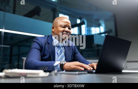 Reifer schwarzer Geschäftsmann lächelt, während er in modernen Büros an einem Laptop arbeitet Stockfoto