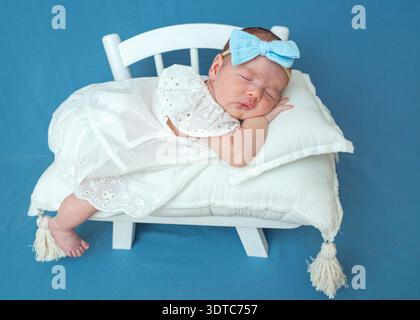 Schlafendes Kind auf weißer Bank mit Kissen vor blauem Hintergrund. Saubere, moderne Komposition, ideal für Babyfotografie und Designkonzepte. Stockfoto