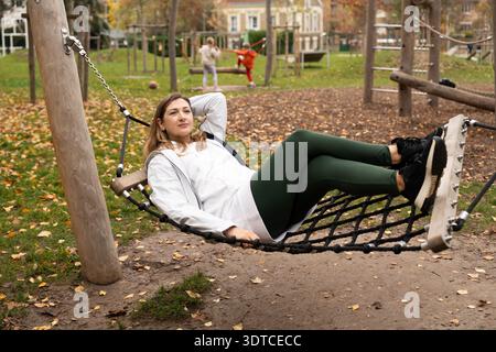 Entspannte Mutter, die auf dem Spielplatz liegt und Kinder in der Nähe beobachtet Stockfoto