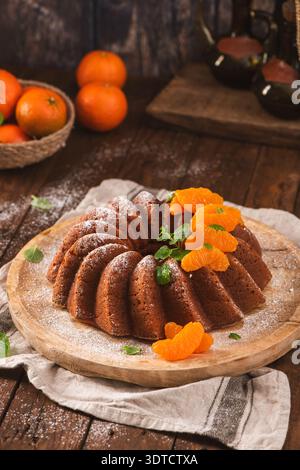 Ein feuchter Zitruskuchen sitzt auf einer rustikalen Holzplatte, bestäubt mit Puderzucker und gekrönt mit lebhaften Mandarinensegmenten und Minzblättern im Krieg Stockfoto
