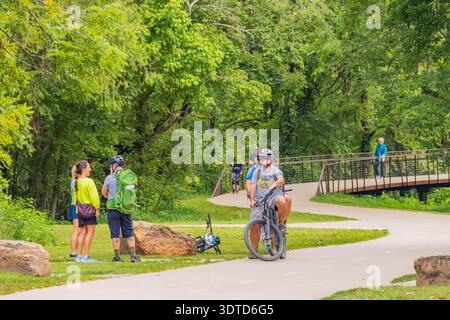 Arkansas, 10. August 2024 – Radfahrer und Wanderer treffen sich auf einem gewundenen, gepflasterten Weg, umgeben von dichtem Grün und einer Holzbrücke in der Ferne. Stockfoto