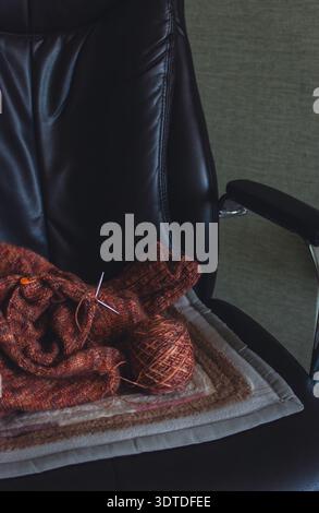 Unfertiger, melierter brauner Pullover mit Stricknadeln am Sessel. Stricknadeln und Kugeln aus warmem, meliertem Garn. Strickarbeit läuft. Stockfoto