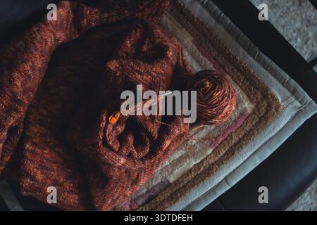 Unfertiger, melierter brauner Pullover mit Stricknadeln am Sessel. Stricknadeln und Kugeln aus warmem, meliertem Garn. Strickarbeit läuft. Stockfoto