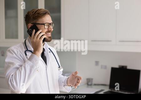 Lächelnder junger Arzt, der aus dem Klinikbüro telefoniert Stockfoto