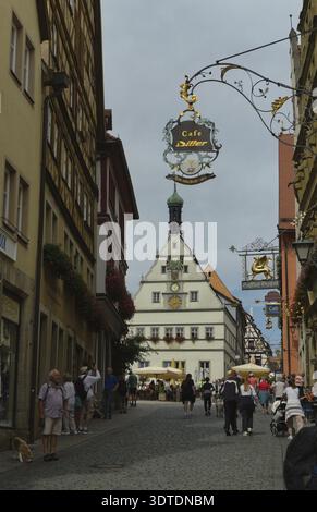 Eine kopfsteingepflasterte Straße, gesäumt von historischen Gebäuden und hängenden Schildern, wo Menschen an einem bewölkten Tag in Rothenburg Tauber draußen spazieren und sitzen Stockfoto