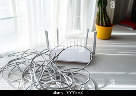 Moderner Wireless-Router auf einem Tisch mit einem Haufen grauer Netzwerk-Patchkabel in einem sonnigen Innenraum. Konzept der Installation des Heimnetzwerks, Kabelmanagement, Stockfoto