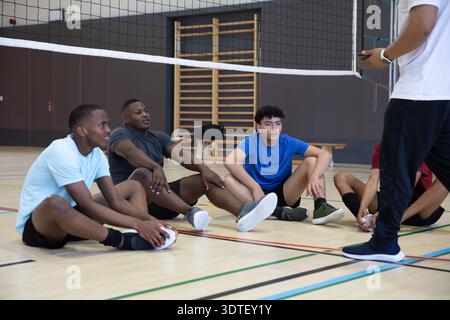 Verschiedene männliche Mannschaftskameraden sitzen auf dem Fitnessraum unter dem Volleyballnetz und lauschen dem Trainer mit Stoppuhr Stockfoto