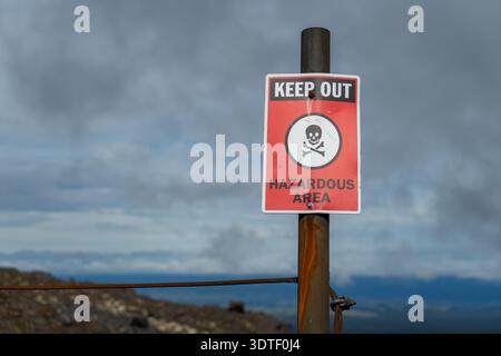 Halten Sie das Gefahrenzeichen fern. Auffälliges rot-schwarz-weißes Schild mit grafischem Symbol eines Schädels und gekreuzter Knochen. Hintergrund von felsigem Hang und bewölktem Himmel. Stockfoto