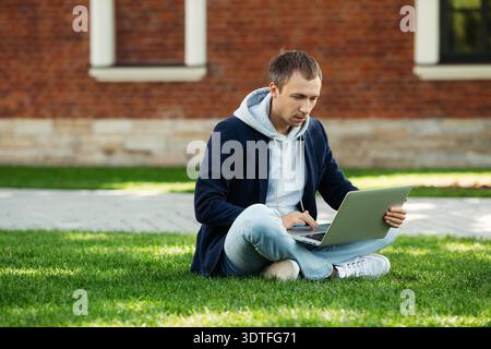 Konzentrierter junger Programmierer, der im Park mit überkreuzten Beinen auf Gras sitzt und sich auf die Prüfungen vorbereitet. Freischaffender Blogger männlich in Freizeitkleidung, der außerhalb auf dem Rasen am Computer arbeitet. || Modell freigegeben Stockfoto