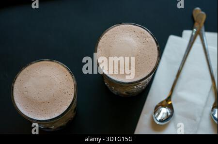 Blick von oben auf zwei Gläser cremigen heißen Kakao mit glattem Milchschaum, serviert mit Löffeln und Serviette. Gemütliche Café-Atmosphäre und warmes Getränk. Stockfoto