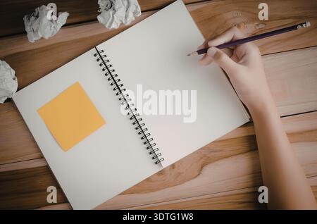 Eine Hand, die einen Bleistift über ein Notizbuch hält, mit einer Haftnotiz auf einem Holztisch Stockfoto