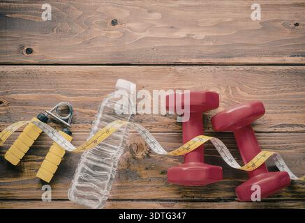 Ein Foto von Fitnessgeräten, einschließlich Kurzhanteln, Maßband, Sprungseil und Wasserflasche auf hölzernem Hintergrund. Stockfoto