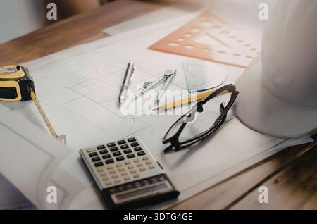 Eine Sammlung von Werkzeugen für die Bauplanung, darunter ein Taschenrechner, eine Brille, Bleistifte und ein Schutzhelm, auf einem Schreibtisch mit Blaupausen. Stockfoto