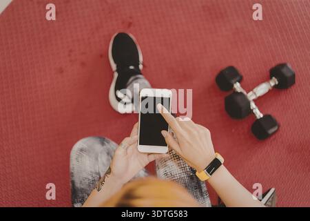 Eine Frau sitzt auf einer roten Yogamatte und macht eine Pause von ihrem Workout, um ihr Handy zu überprüfen. Sie trägt eine schwarze Smartwatch und ein orangefarbenes Band am Handgelenk. Stockfoto