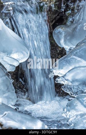 Frosted Waterfall Cascade: Eiskalte Skulpturen umschließen einen kleinen Wasserfall, während das Wasser in einer ruhigen Winterlandschaft hindurchstürzt. Das Wasser ist teilweise f Stockfoto