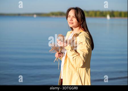 Frau in einem gelben Regenmantel, die getrocknete Blumen am See hält und die warmen Sonnenstrahlen genießt Stockfoto