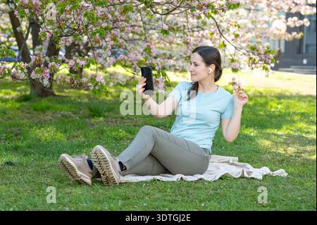 Eine junge Frau sitzt auf einer Decke in einem blühenden Frühlingspark und spricht per Smartphone über einen Videoanruf Stockfoto