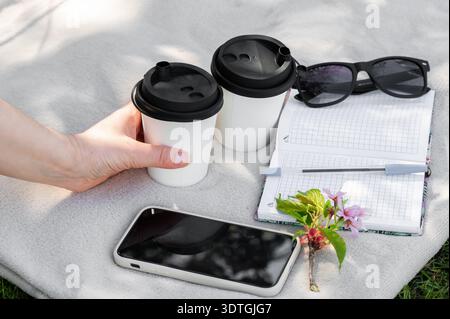 Hand reicht für Papierbecher mit Kaffee oder Tee, auf hellem Hintergrund liegt Notebook, Smartphone und Sakura Zweig, freiberuflich in der Natur Stockfoto