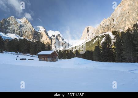 Schneebedeckte Almhütte im Fassatal mit Dolomitengipfeln und Kiefernwäldern, Trentino Südtirol, Italien Stockfoto