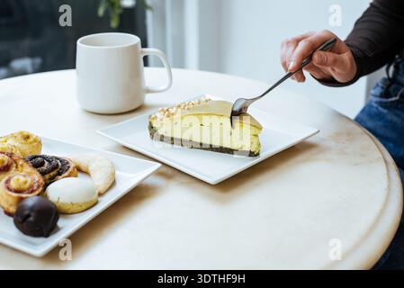 Eine Person sitzt mit einer Tasse Kaffee an einem kleinen Tisch. Sie essen gleich eine Scheibe Käsekuchen. Ein Teller mit verschiedenen Gebäckstücken steht in der Nähe und kreiert Stockfoto
