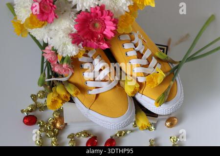 Gelbe Sneakers mit gelben Narzissen und Schokoladeneiern auf rosafarbenem Hintergrund mit Kopierraum. Osterfest-Konzept. Stockfoto