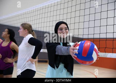 Diverse Teenager-Volleyballmannschaft, die im Fitnessstudio trainiert, Hijab-Spieler, die rot, weiß oder blau halten Stockfoto
