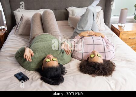 Diverse Freundinnen liegen nebeneinander auf dem Bett im Schlafzimmer mit Gurkenscheiben, Smartphone in der Nähe Stockfoto