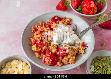 Erdbeercrumble-Dessert mit schmelzendem Vanilleeis, Haferbelag, Kürbiskernen und Mandeln in einer Schüssel auf rosa Hintergrund. Hausgemachtes süßes Gebäck Stockfoto